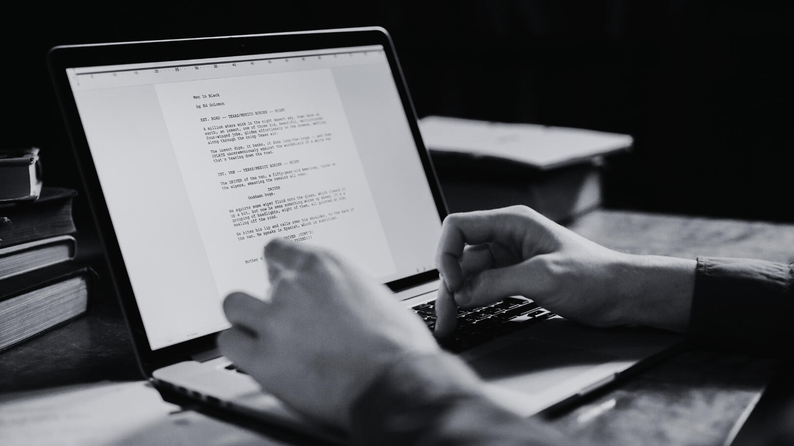 Screenwriting & Concept Development - Jessica Lima Primo piano in bianco e nero di mani che scrivono una sceneggiatura tecnica su un laptop, con lo schermo acceso e dettagli di libri sullo sfondo sfocato.