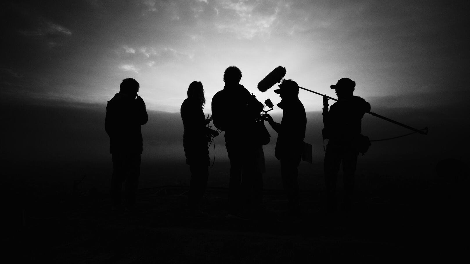 Leadership e Direzione di Troupe — Coordinamento Audiovisivo Un gruppo di lavoro della troupe cinematografica concentrato a esaminare le riprese al monitor sotto fasci di luce da set.