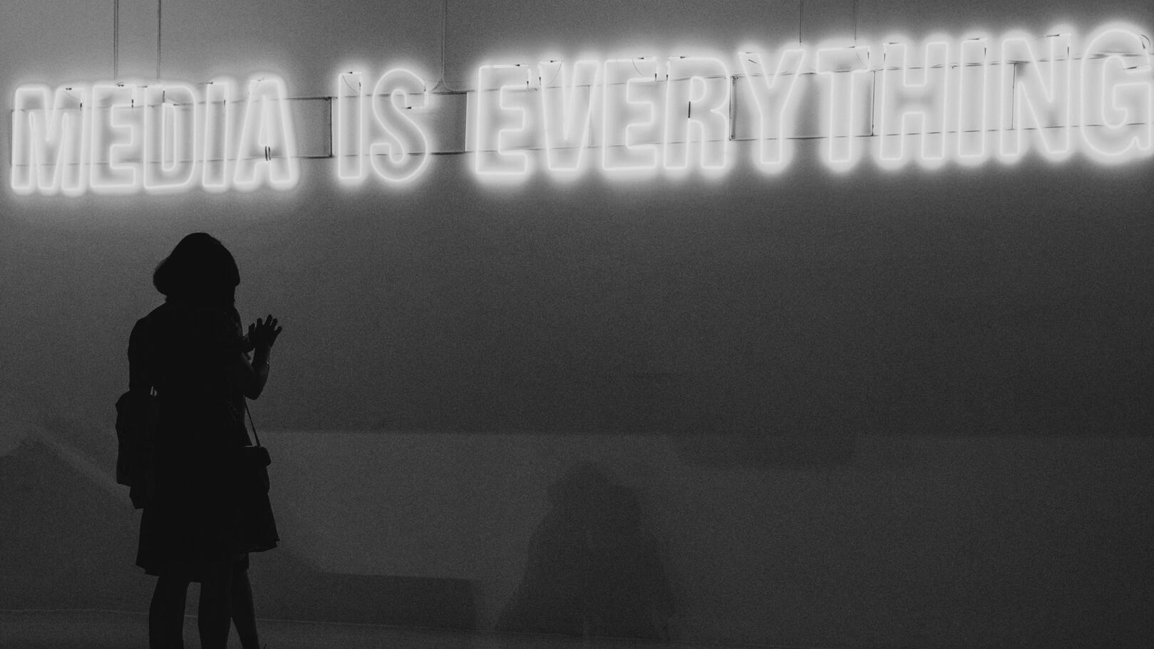 Promotion & Media Planning — La Strategia Media e Diffusione dei Contenuti Una silhouette di persona di spalle osserva una scritta al neon luminosa che recita: "MEDIA IS EVERYTHING".